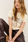 A woman wearing a cream-colored graphic t-shirt with 'BASH printed on the front, paired with dark brown pants, sitting against a plain off-white background.