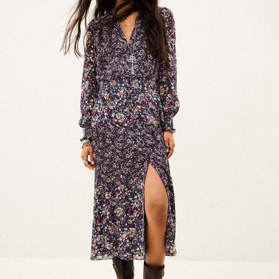 A woman modeling a long-sleeved midi dress with a dense multicolor floral print on a dark base, featuring a high thigh slit and a V-neckline, paired with black boots against a plain white studio background.