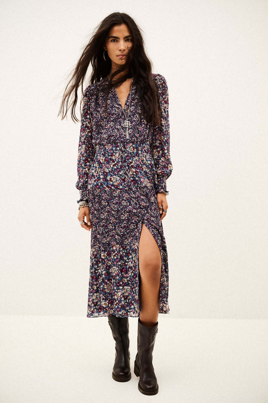 A woman modeling a long-sleeved midi dress with a dense multicolor floral print on a dark base, featuring a high thigh slit and a V-neckline, paired with black boots against a plain white studio background.