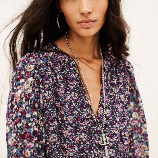 A woman wearing a long-sleeve floral print top with a deep V-neckline, layered with silver necklaces, against a plain off-white background.