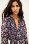 A woman wearing a long-sleeve floral print top with a deep V-neckline, layered with silver necklaces, against a plain off-white background.