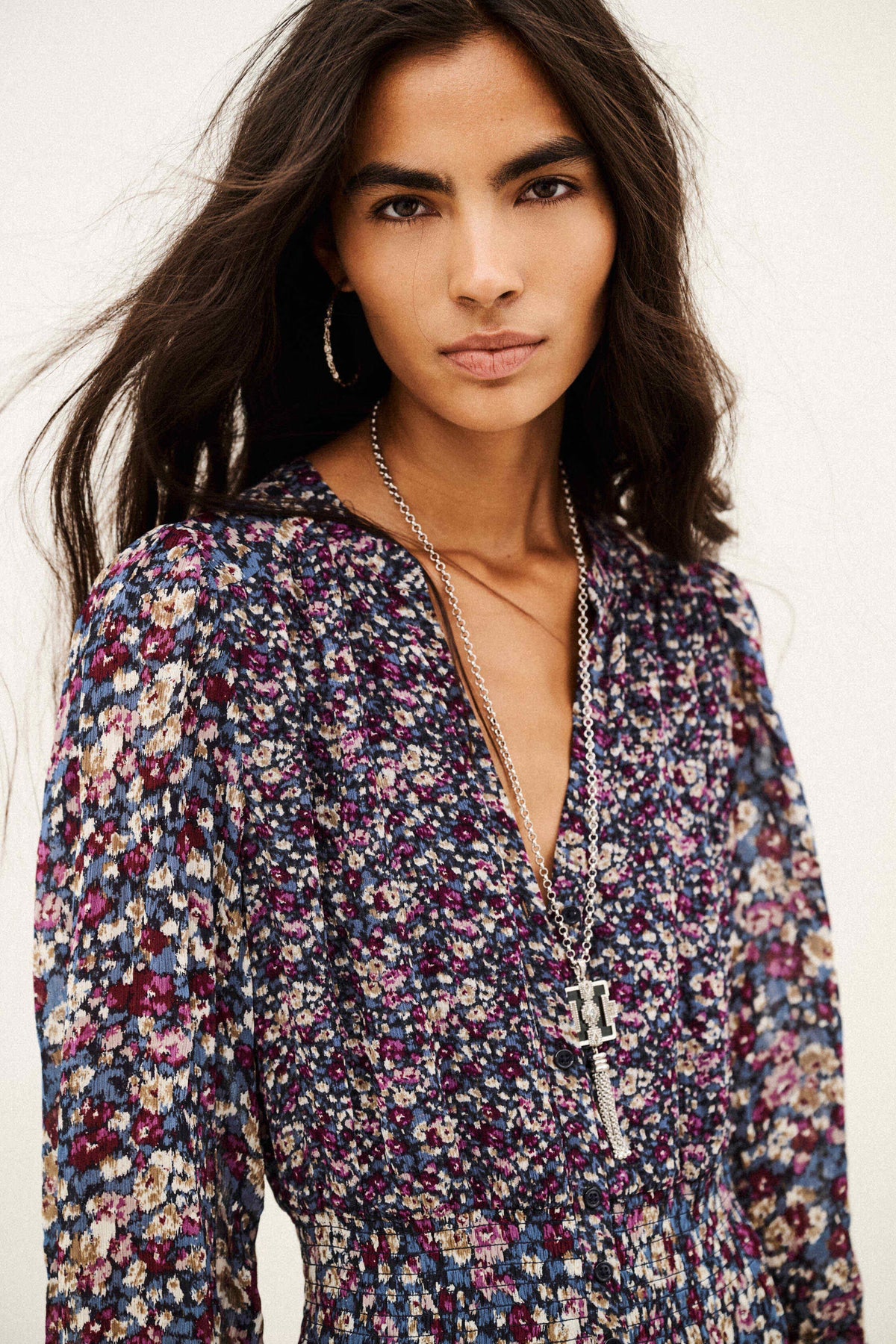 A woman wearing a long-sleeve floral print top with a deep V-neckline, layered with silver necklaces, against a plain off-white background.