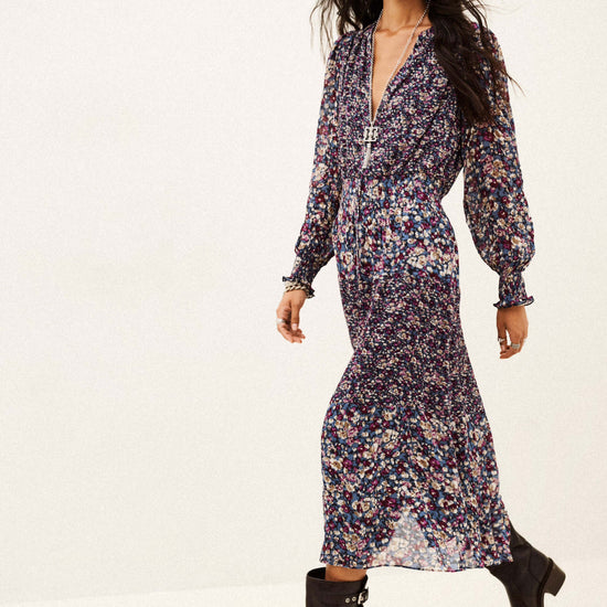 A woman modeling a long-sleeved, deep V-neck midi dress with a dense multicolor floral print in purple, blue, pink, and green on a dark base, paired with black ankle boots, against a plain white studio background.
