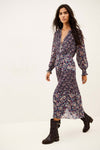 A woman modeling a long-sleeved, deep V-neck midi dress with a dense multicolor floral print in purple, blue, pink, and green on a dark base, paired with black ankle boots, against a plain white studio background.