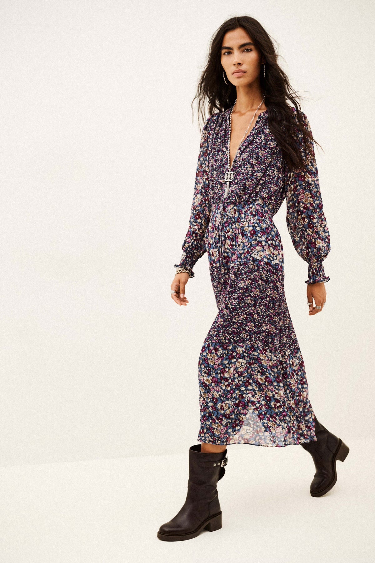 A woman modeling a long-sleeved, deep V-neck midi dress with a dense multicolor floral print in purple, blue, pink, and green on a dark base, paired with black ankle boots, against a plain white studio background.