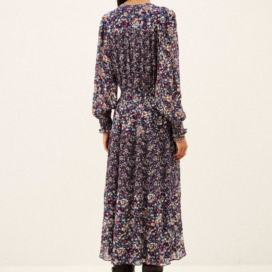 A woman viewed from the back wearing a long-sleeve midi dress with a dense multicolor floral print on a dark base, paired with black ankle boots, against a plain off-white studio background.
