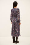 A woman viewed from the back wearing a long-sleeve midi dress with a dense multicolor floral print on a dark base, paired with black ankle boots, against a plain off-white studio background.