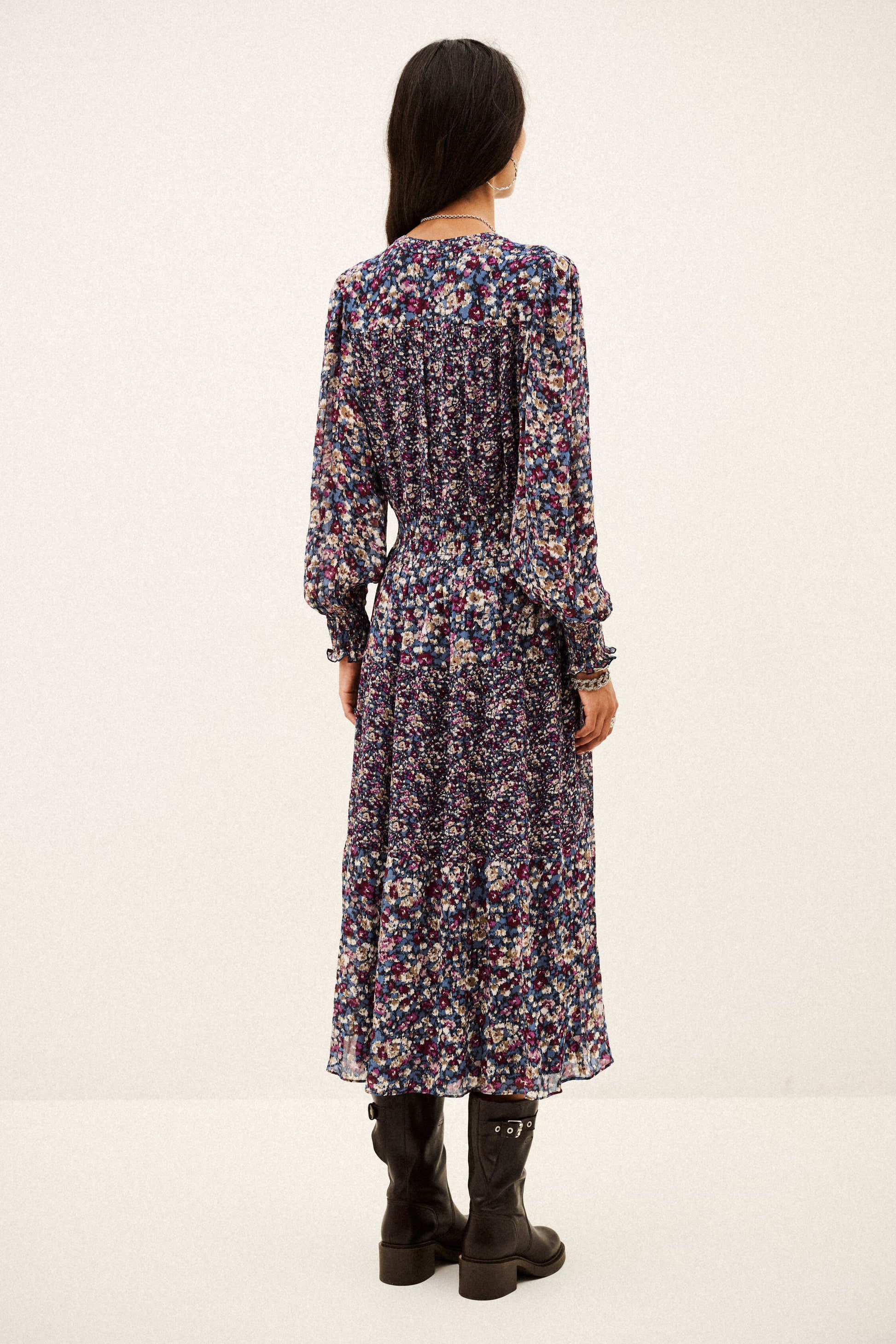 A woman viewed from the back wearing a long-sleeve midi dress with a dense multicolor floral print on a dark base, paired with black ankle boots, against a plain off-white studio background.