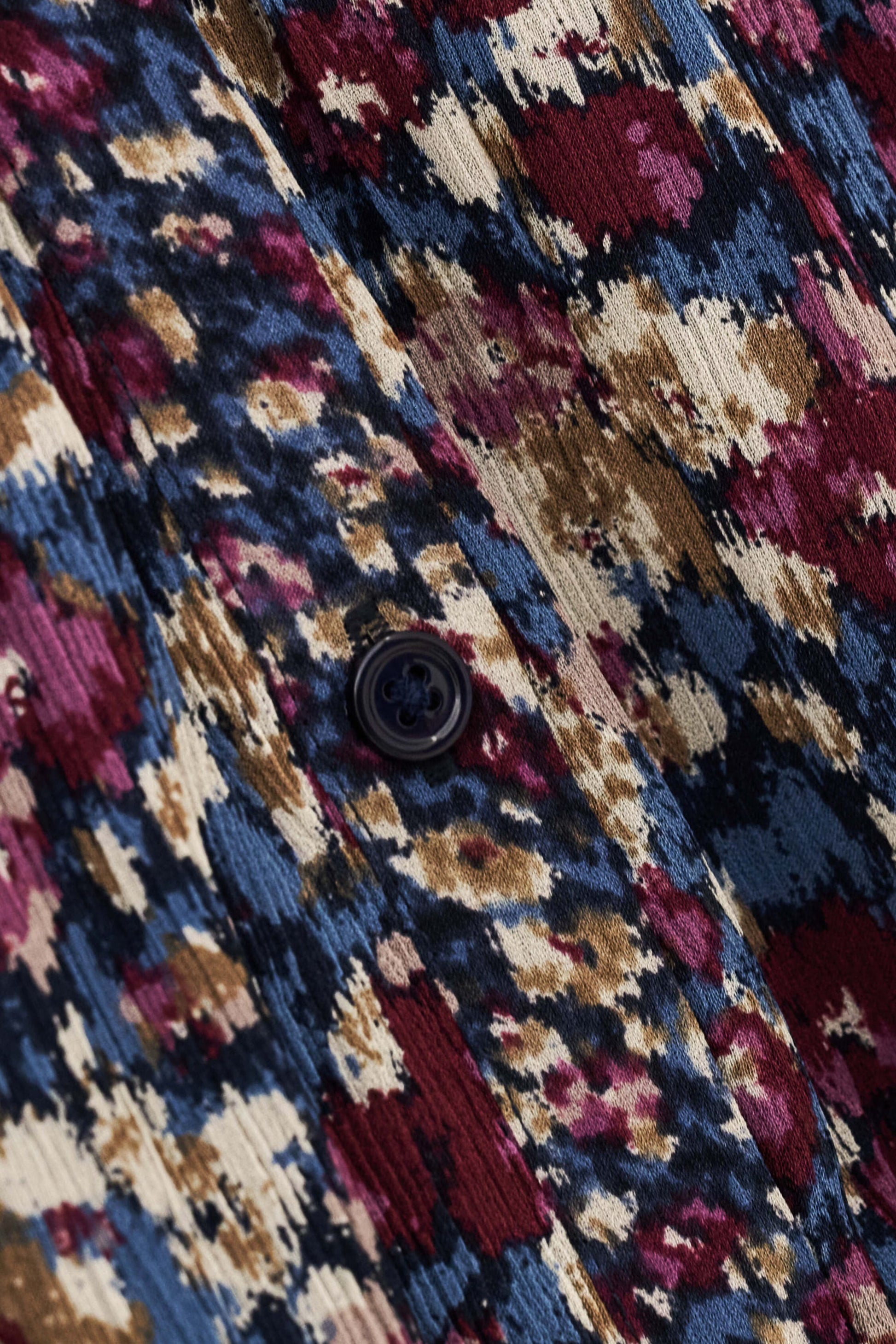 Close-up of a textured fabric with an abstract ikat pattern in navy blue, burgundy, beige, and brown, featuring a dark button.