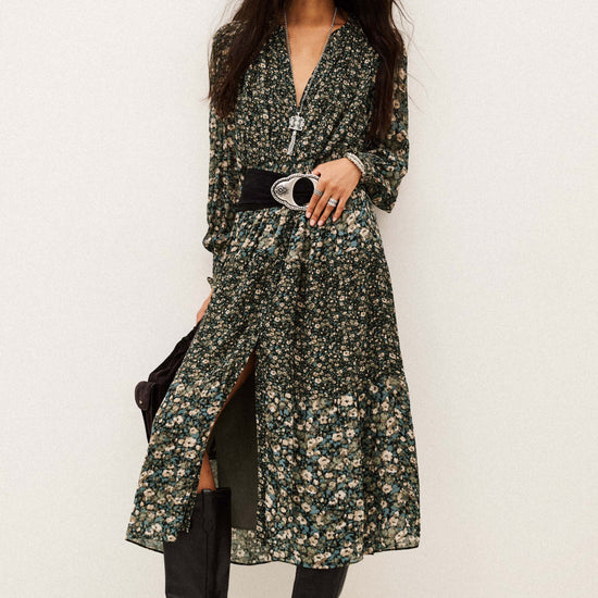 A model wearing a long-sleeved, dark green floral print midi dress with a black belt and black knee-high boots, standing against a plain off-white studio background.