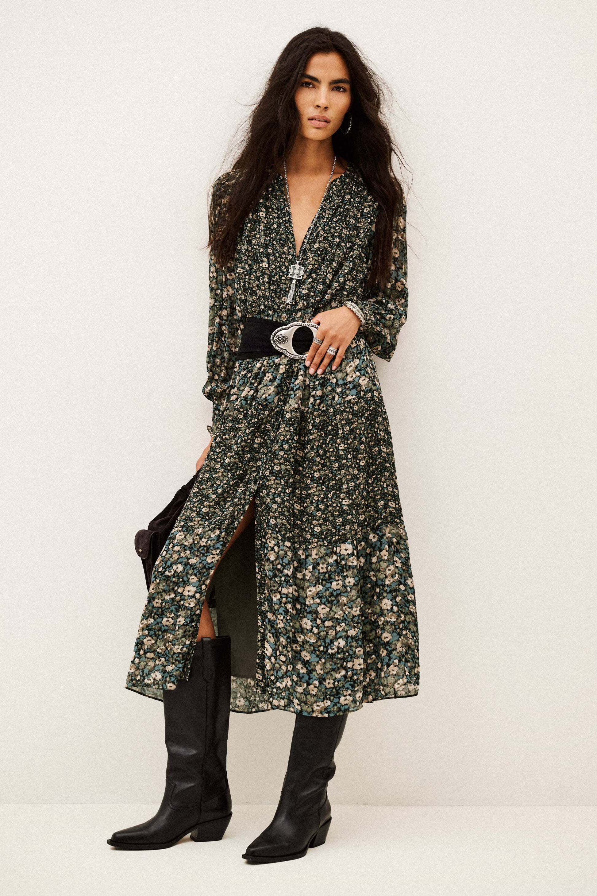 A model wearing a long-sleeved, dark green floral print midi dress with a black belt and black knee-high boots, standing against a plain off-white studio background.