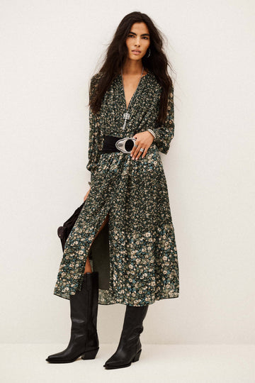 A model wearing a long-sleeved, dark green floral print midi dress with a black belt and black knee-high boots, standing against a plain off-white studio background.