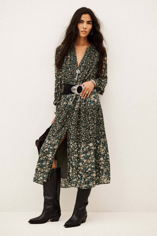 A model wearing a long-sleeved, dark green floral print midi dress with a black belt and black knee-high boots, standing against a plain off-white studio background.