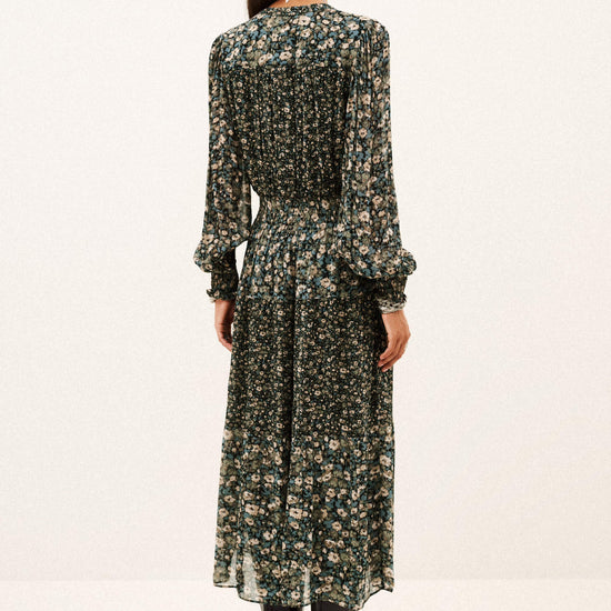 A woman viewed from the back wearing a long-sleeve midi dress with a dense dark green and beige floral print, paired with black ankle boots, against a plain white studio background.