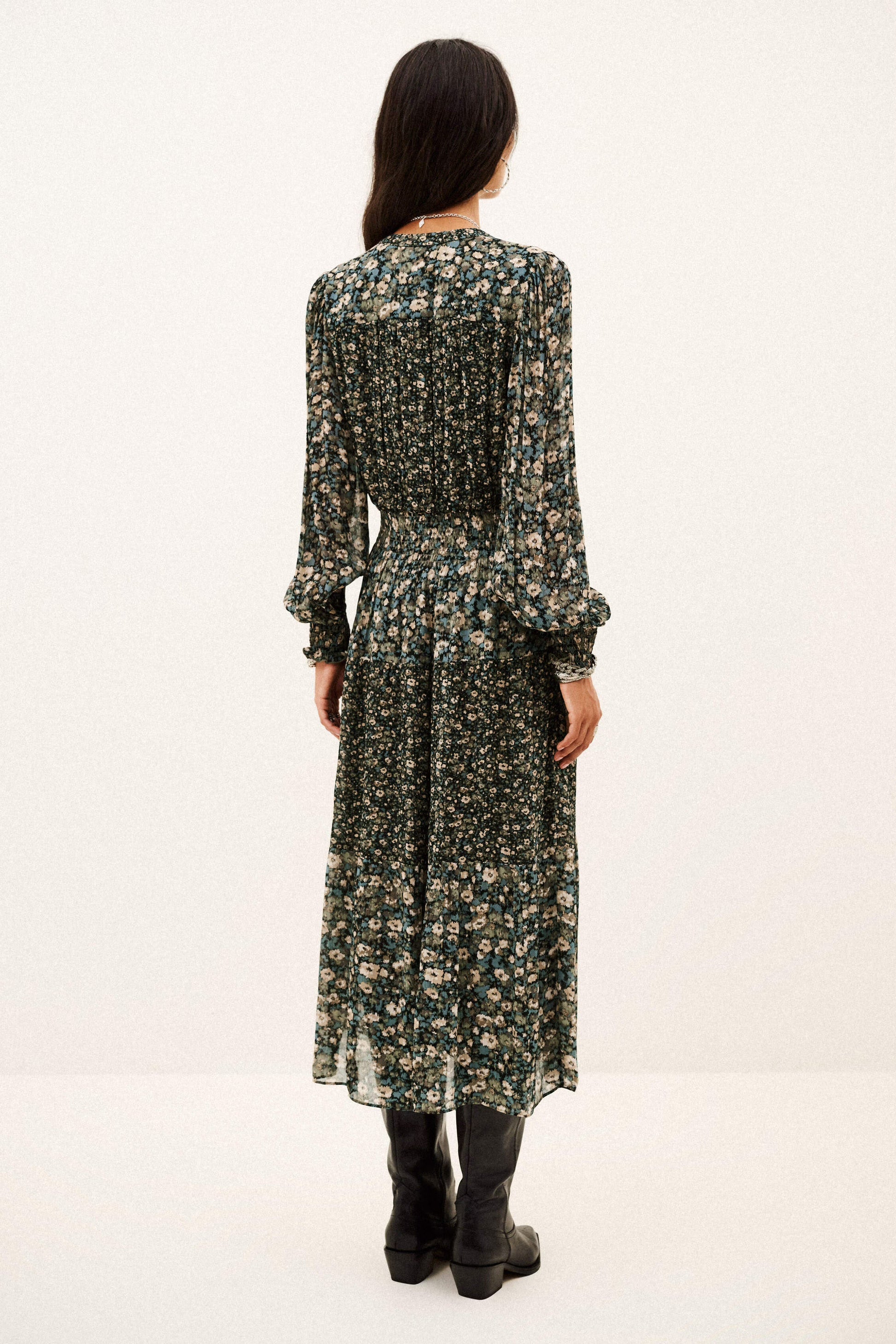 A woman viewed from the back wearing a long-sleeve midi dress with a dense dark green and beige floral print, paired with black ankle boots, against a plain white studio background.