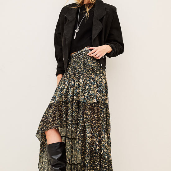 A model stands against a plain light beige studio background wearing a long black jacket, a high-low floral skirt with a black base and muted green and gold floral print, and black knee-high heeled boots.