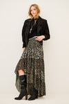 A model stands against a plain light beige studio background wearing a long black jacket, a high-low floral skirt with a black base and muted green and gold floral print, and black knee-high heeled boots.