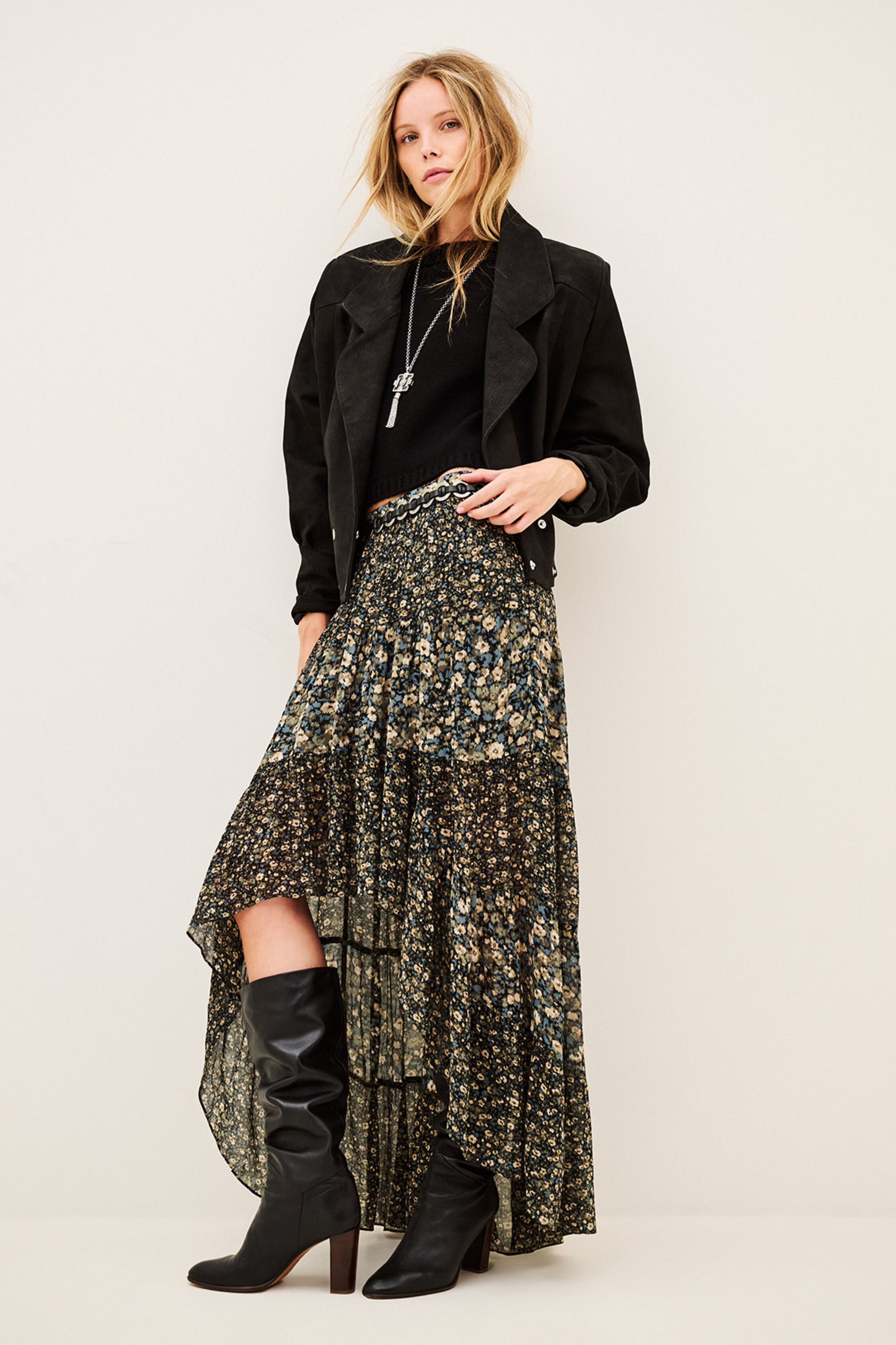 A model stands against a plain light beige studio background wearing a long black jacket, a high-low floral skirt with a black base and muted green and gold floral print, and black knee-high heeled boots.