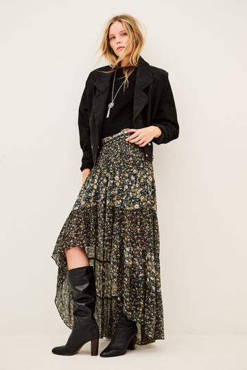 A model stands against a plain light beige studio background wearing a long black jacket, a high-low floral skirt with a black base and muted green and gold floral print, and black knee-high heeled boots.
