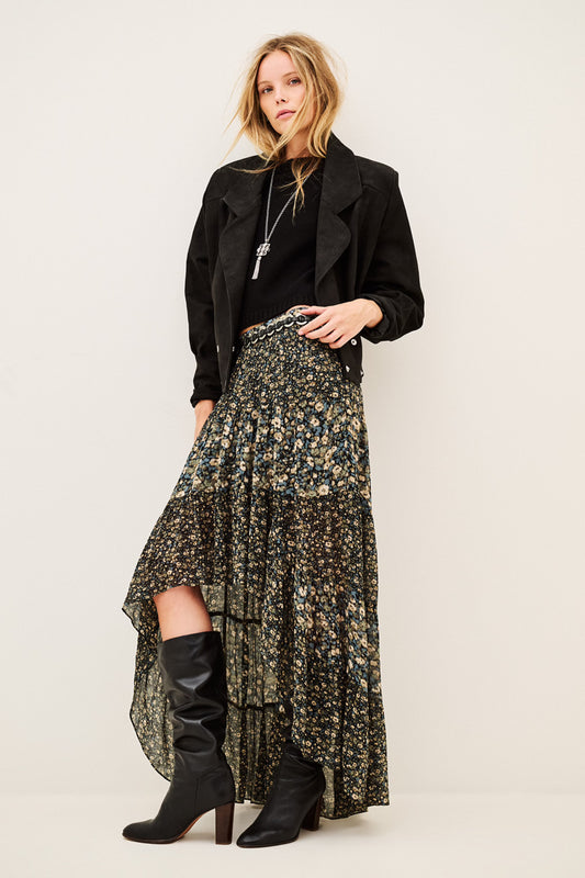 A model stands against a plain light beige studio background wearing a long black jacket, a high-low floral skirt with a black base and muted green and gold floral print, and black knee-high heeled boots.