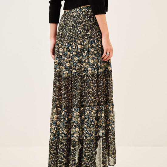 A model from the back wearing a long black floral print skirt with beige and yellow flowers, paired with a black long-sleeve top and black boots, against a plain off-white studio background.