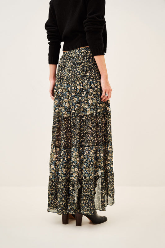 A model from the back wearing a long black floral print skirt with beige and yellow flowers, paired with a black long-sleeve top and black boots, against a plain off-white studio background.