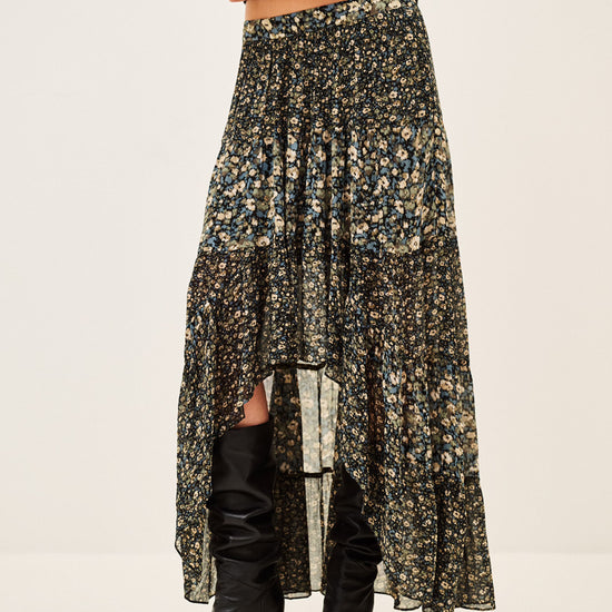 A model wearing a long, high-low floral print skirt in black, olive green, and beige, paired with black knee-high boots against a plain off-white studio background.