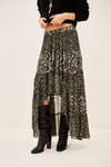 A model wearing a long, high-low floral print skirt in black, olive green, and beige, paired with black knee-high boots against a plain off-white studio background.