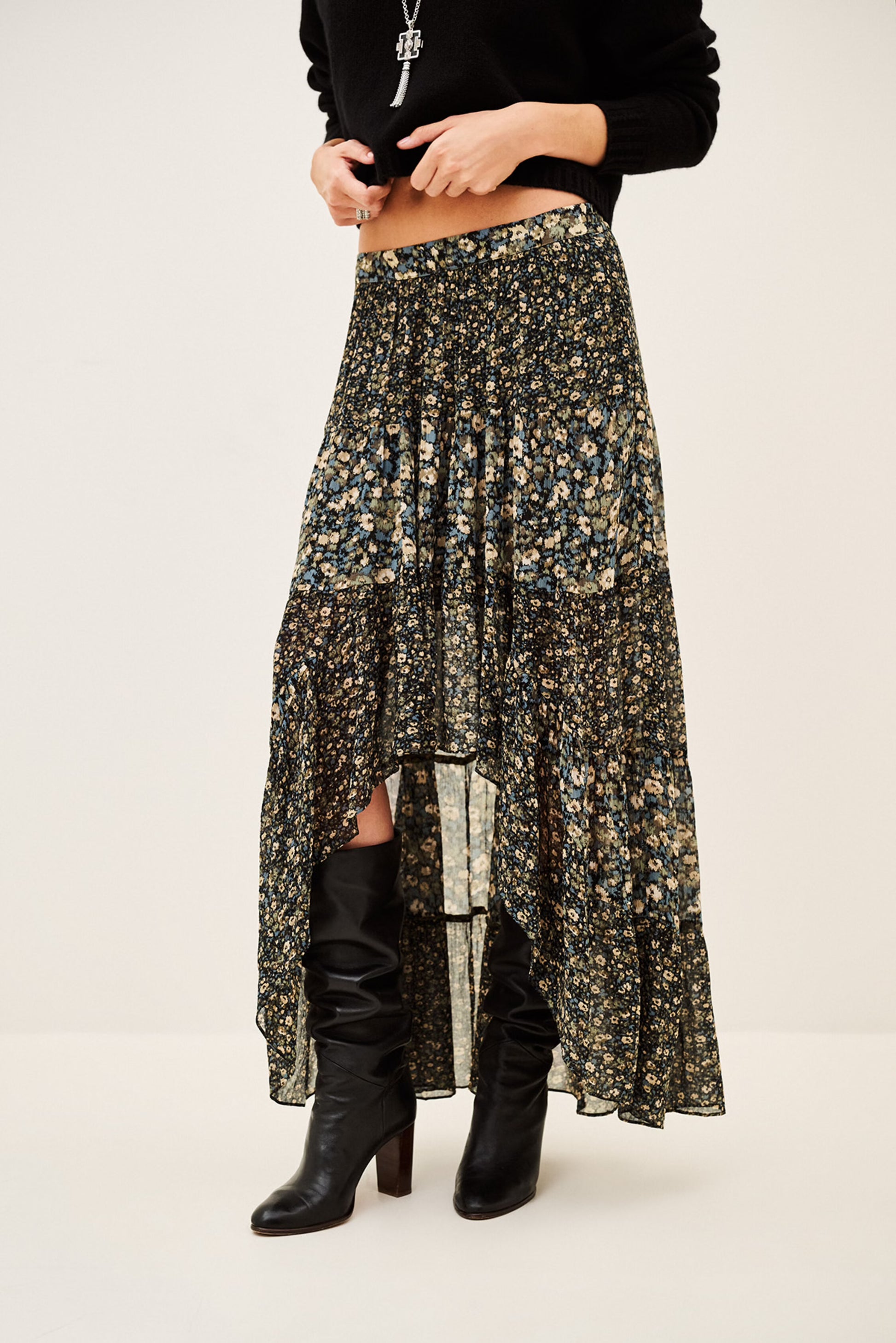 A model wearing a long, high-low floral print skirt in black, olive green, and beige, paired with black knee-high boots against a plain off-white studio background.