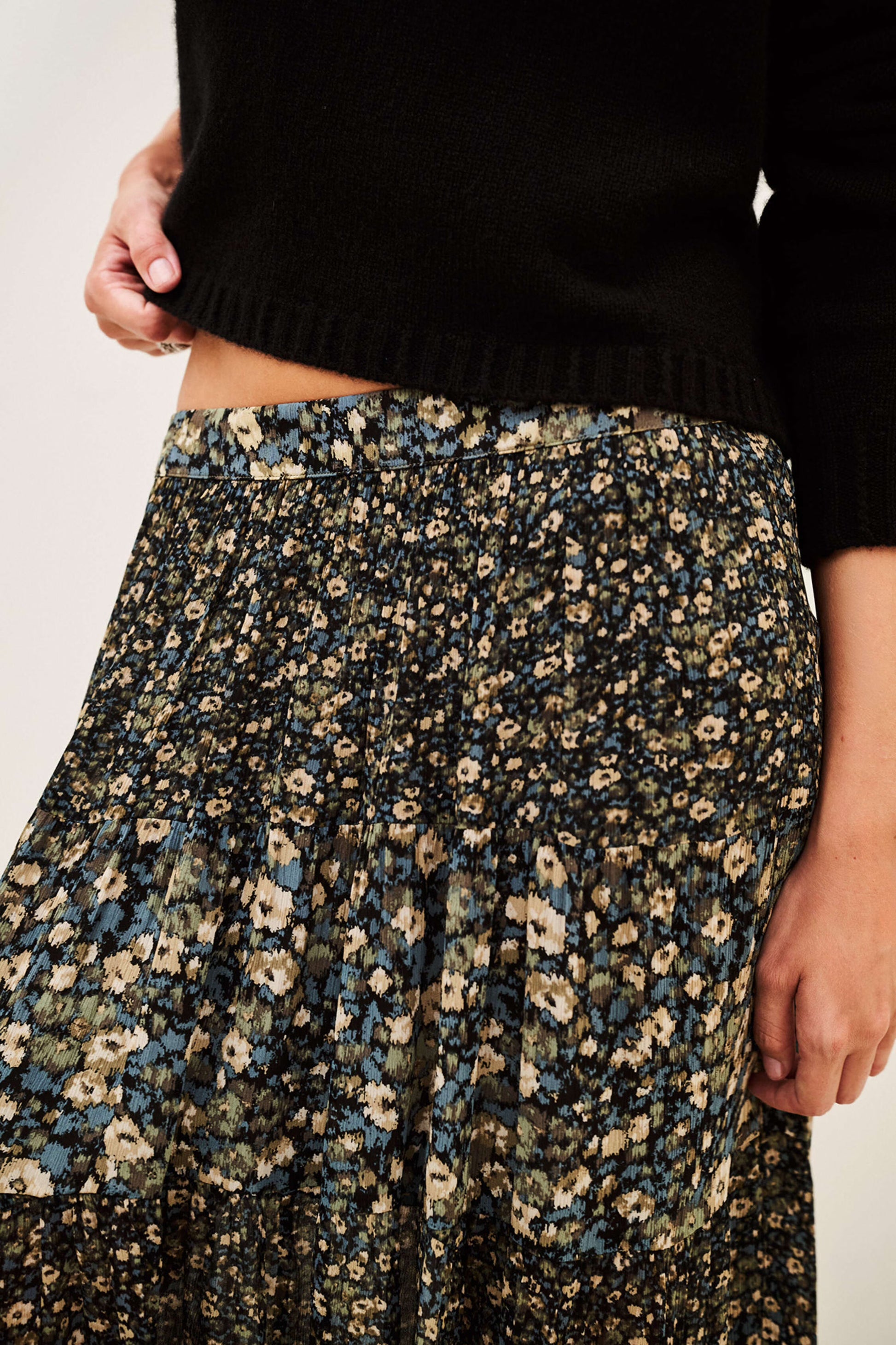 Close-up of a person wearing a black knit sweater and a dark floral skirt with blue, beige, and cream flowers, against a plain light background.