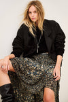 A woman seated against a plain light background wearing a black jacket, black top, a dark floral midi skirt with blue and beige floral details, and black knee-high boots.