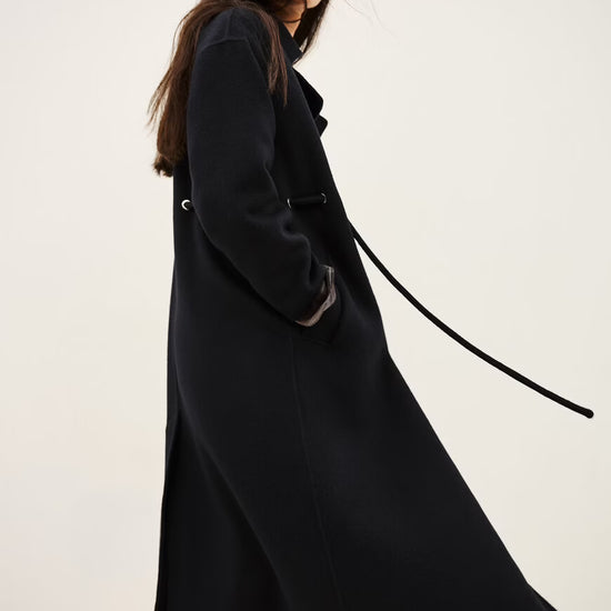 A woman wearing a long black coat with a matching belt, paired with blue jeans and brown boots, photographed against a plain light background.