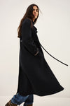 A woman wearing a long black coat with a matching belt, paired with blue jeans and brown boots, photographed against a plain light background.