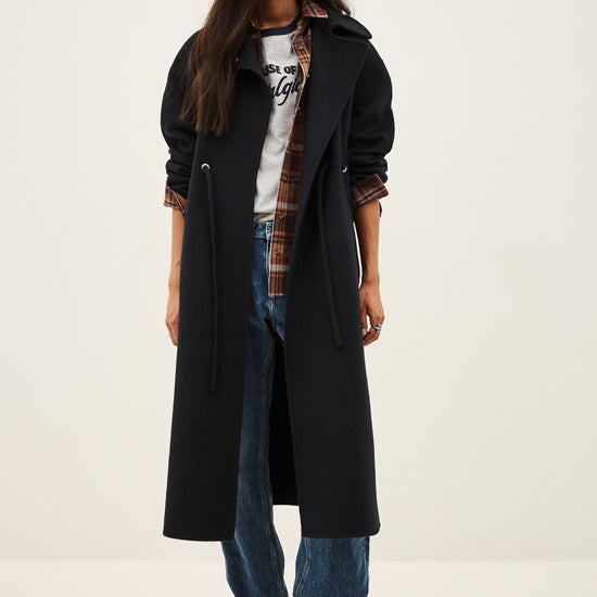 A model stands against a plain light background wearing a long black coat over a white graphic T-shirt, a brown plaid shirt, blue jeans, and brown ankle boots.