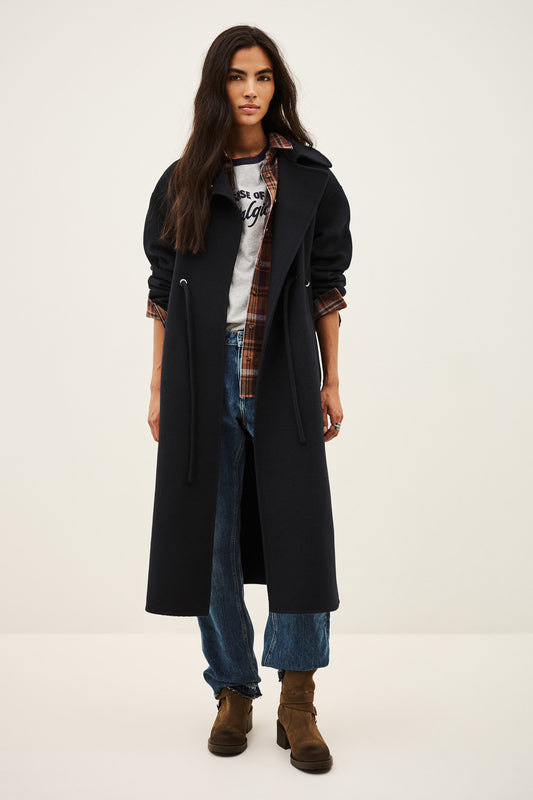 A model stands against a plain light background wearing a long black coat over a white graphic T-shirt, a brown plaid shirt, blue jeans, and brown ankle boots.