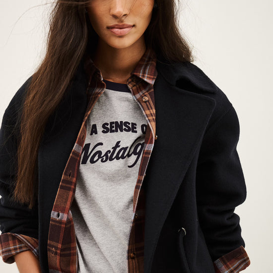 A woman wearing a black coat over a gray graphic t-shirt and a brown plaid flannel shirt, posed against a light off-white background.
