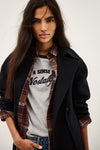 A woman wearing a black coat over a gray graphic t-shirt and a brown plaid flannel shirt, posed against a light off-white background.