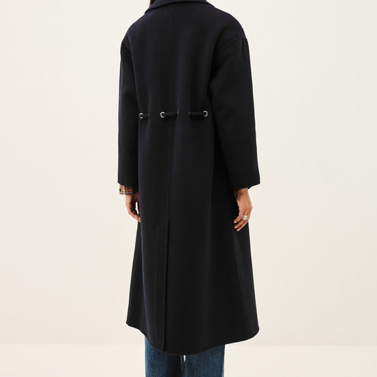 A woman viewed from the back wearing a long black coat with a waist tie, blue jeans, and brown shoes, standing against a plain off-white studio background.
