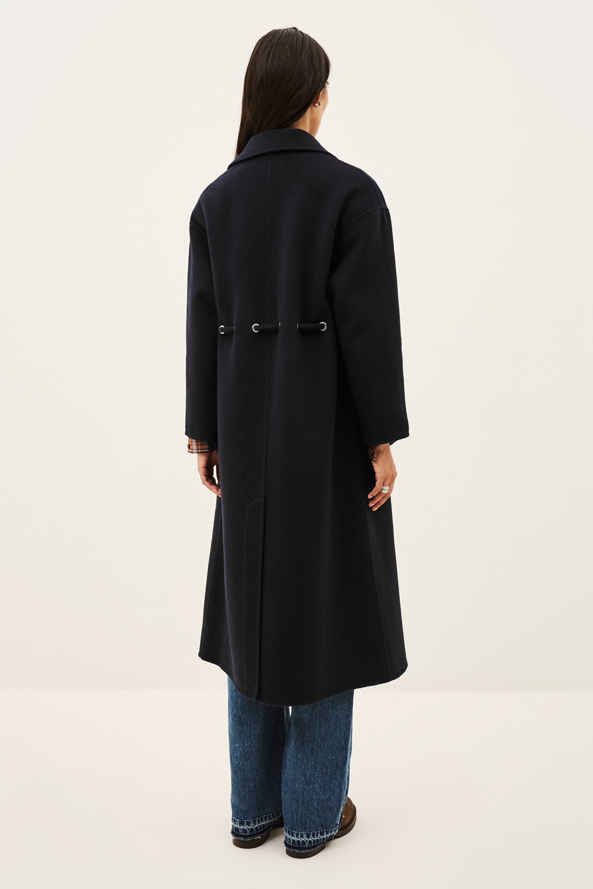 A woman viewed from the back wearing a long black coat with a waist tie, blue jeans, and brown shoes, standing against a plain off-white studio background.