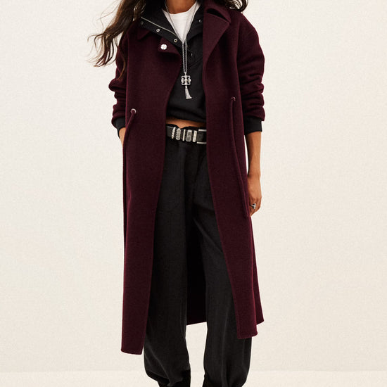 A model wearing a long, dark burgundy wool-blend coat over a black zip-up hoodie, black pants, and black ankle boots, standing against a plain off-white studio background.