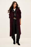 A model wearing a long, dark burgundy wool-blend coat over a black zip-up hoodie, black pants, and black ankle boots, standing against a plain off-white studio background.