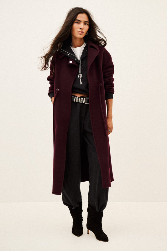 A model wearing a long, dark burgundy wool-blend coat over a black zip-up hoodie, black pants, and black ankle boots, standing against a plain off-white studio background.