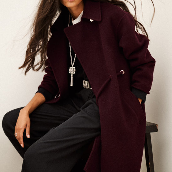 A woman seated on a stool wearing a long burgundy overcoat over a black outfit, photographed against a plain light background.