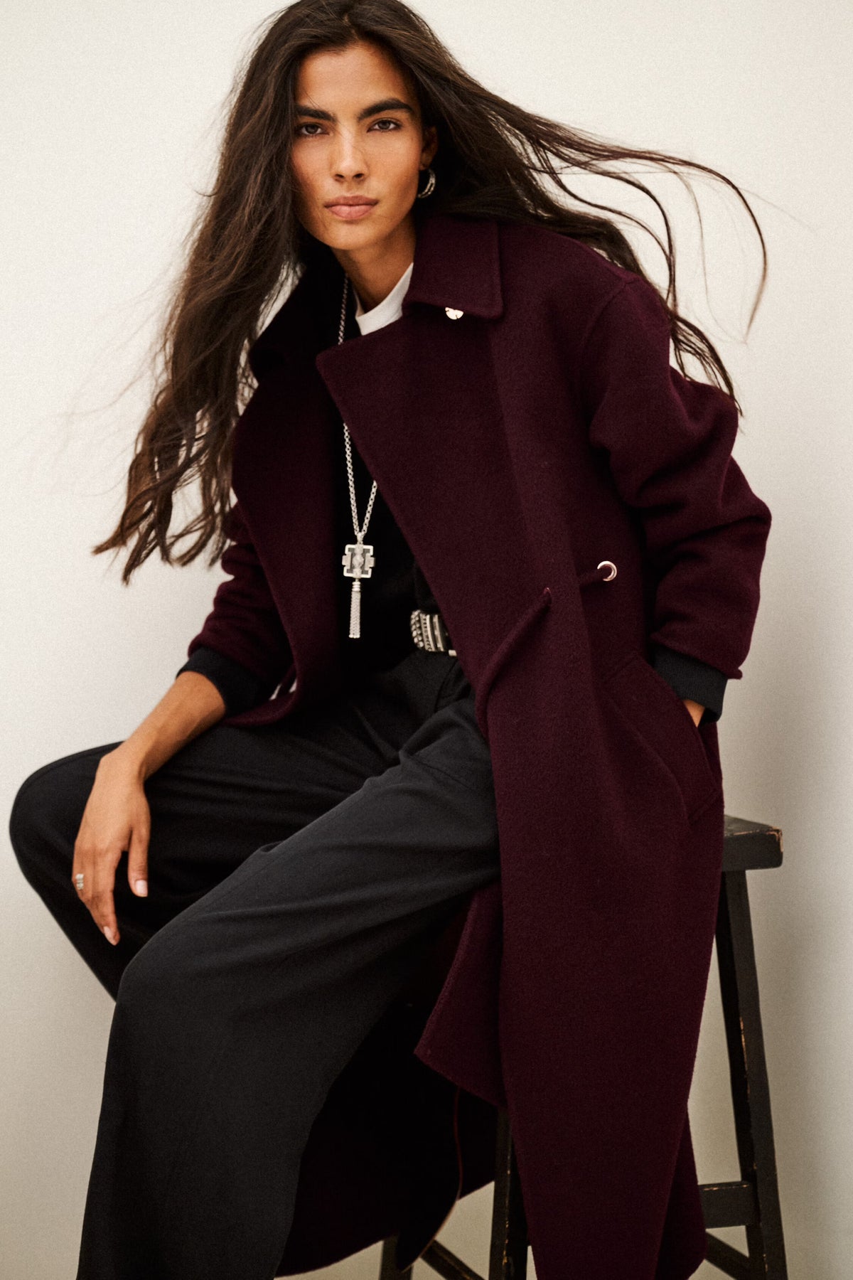 A woman seated on a stool wearing a long burgundy overcoat over a black outfit, photographed against a plain light background.