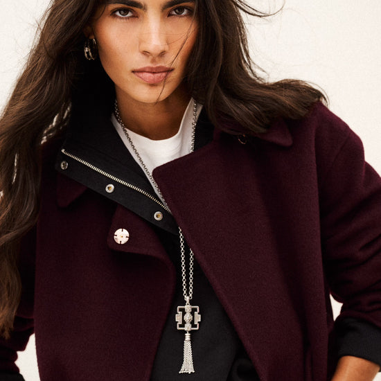 A woman wearing a dark burgundy wool-blend coat over a black top and white undershirt, accessorized with a silver necklace and belt, photographed against a plain off-white background.