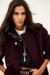 A woman wearing a dark burgundy wool-blend coat over a black top and white undershirt, accessorized with a silver necklace and belt, photographed against a plain off-white background.