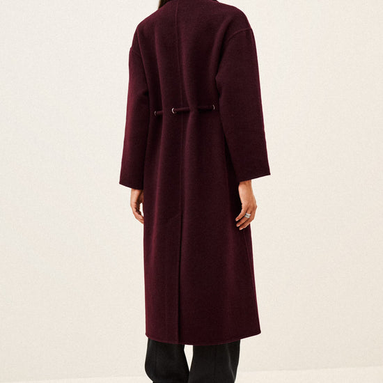 A woman viewed from the back wearing a long burgundy coat with a matching belt, black pants, and black heeled boots against a plain off-white studio background.