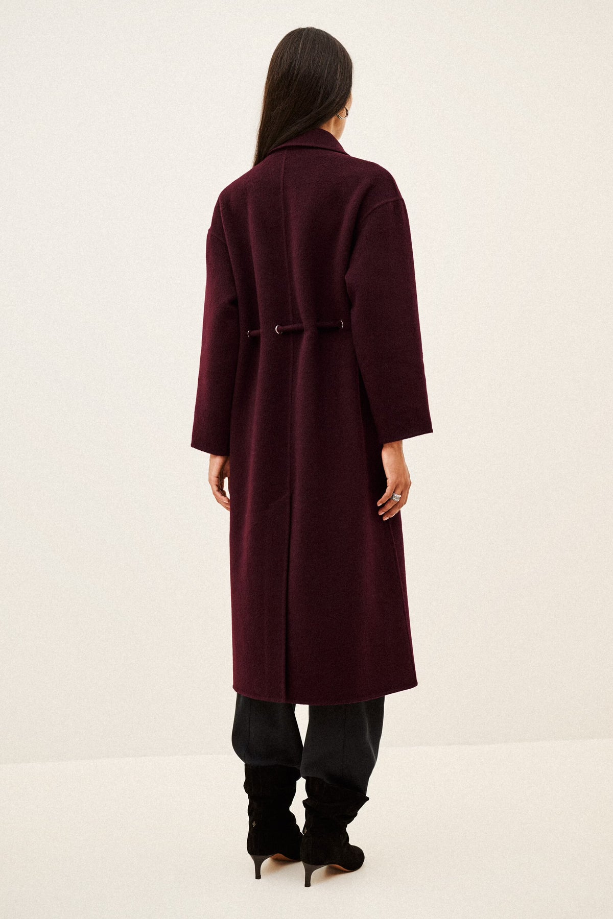 A woman viewed from the back wearing a long burgundy coat with a matching belt, black pants, and black heeled boots against a plain off-white studio background.