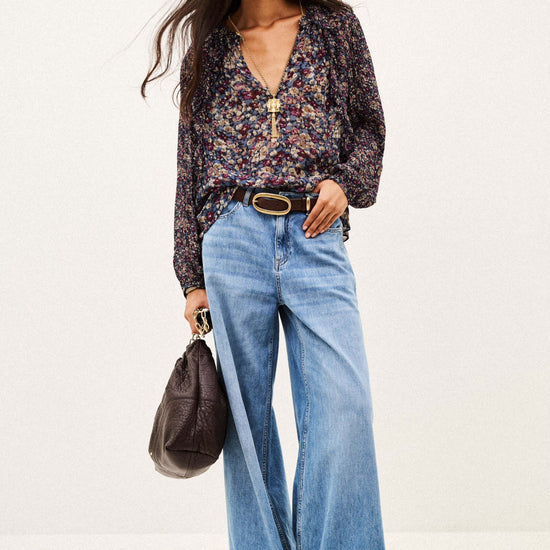 A woman modeling a dark floral long-sleeve blouse tucked into wide-leg light blue jeans, styled with a gold-tone belt, a dark brown handbag, and brown pointed shoes against a plain white studio background.