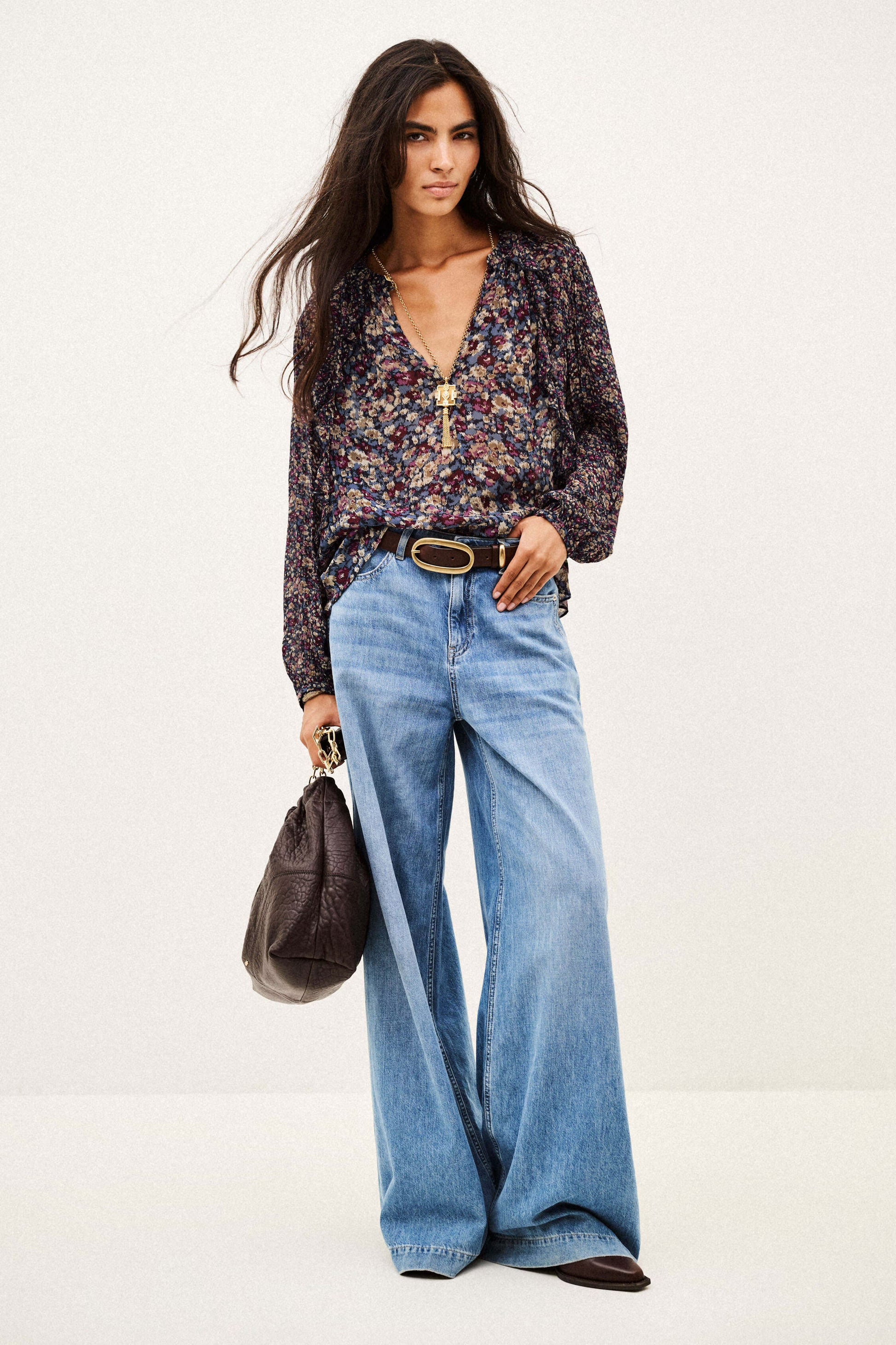 A woman modeling a dark floral long-sleeve blouse tucked into wide-leg light blue jeans, styled with a gold-tone belt, a dark brown handbag, and brown pointed shoes against a plain white studio background.